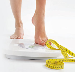 Photo of a women's feet as she steps on a weight scale. A tape measure is positioned on the upper side of the scale.